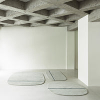Abstract light gray rugs with soft curves and subtle blue lines, set against a minimalist, neutral backdrop.