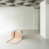 Modern lounge chair in soft pastel pink with a bold maroon stripe, set against a minimalist concrete backdrop.