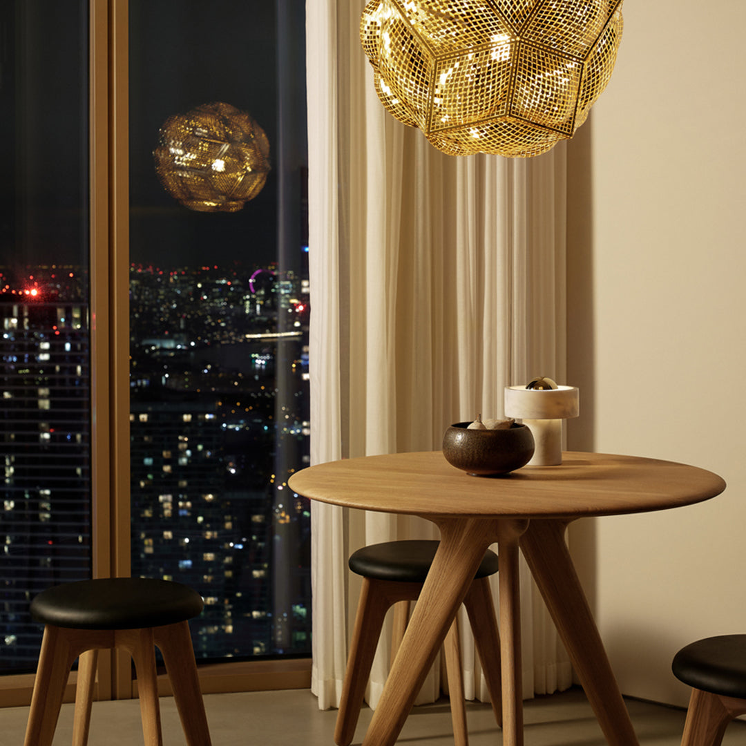 Modern wooden table with black stools, golden pendant light, and soft beige curtains in a chic evening setting.