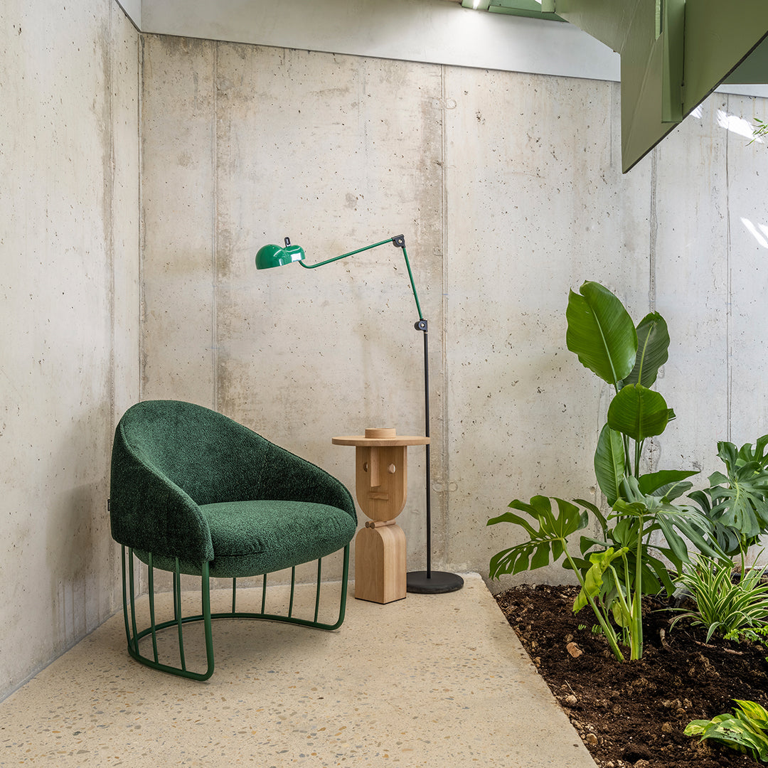 Green plush chair with a curved design, paired with a wooden side table and modern floor lamp. Neutral concrete backdrop.