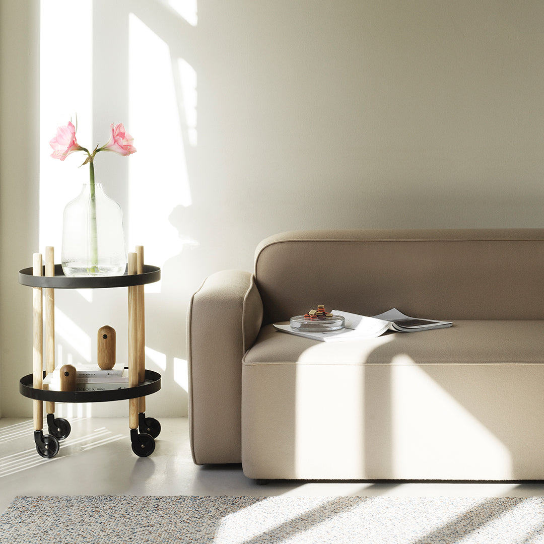 Beige sofa with light wood and black rolling cart, featuring a green vase and pink flowers in a bright, sunlit room.