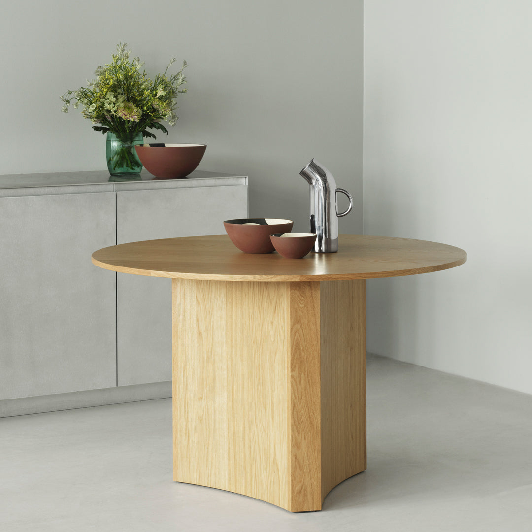 Round wooden dining table with a natural finish and minimalist design, paired with earthy clay bowls and a vase.
