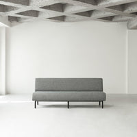 Gray fabric sofa on black legs in a minimalist setting with a textured concrete ceiling and white walls.