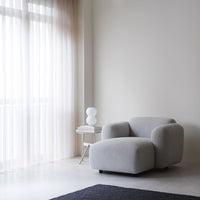 Soft grey upholstered armchair with a minimalist design, set against a neutral backdrop and sheer light curtains.