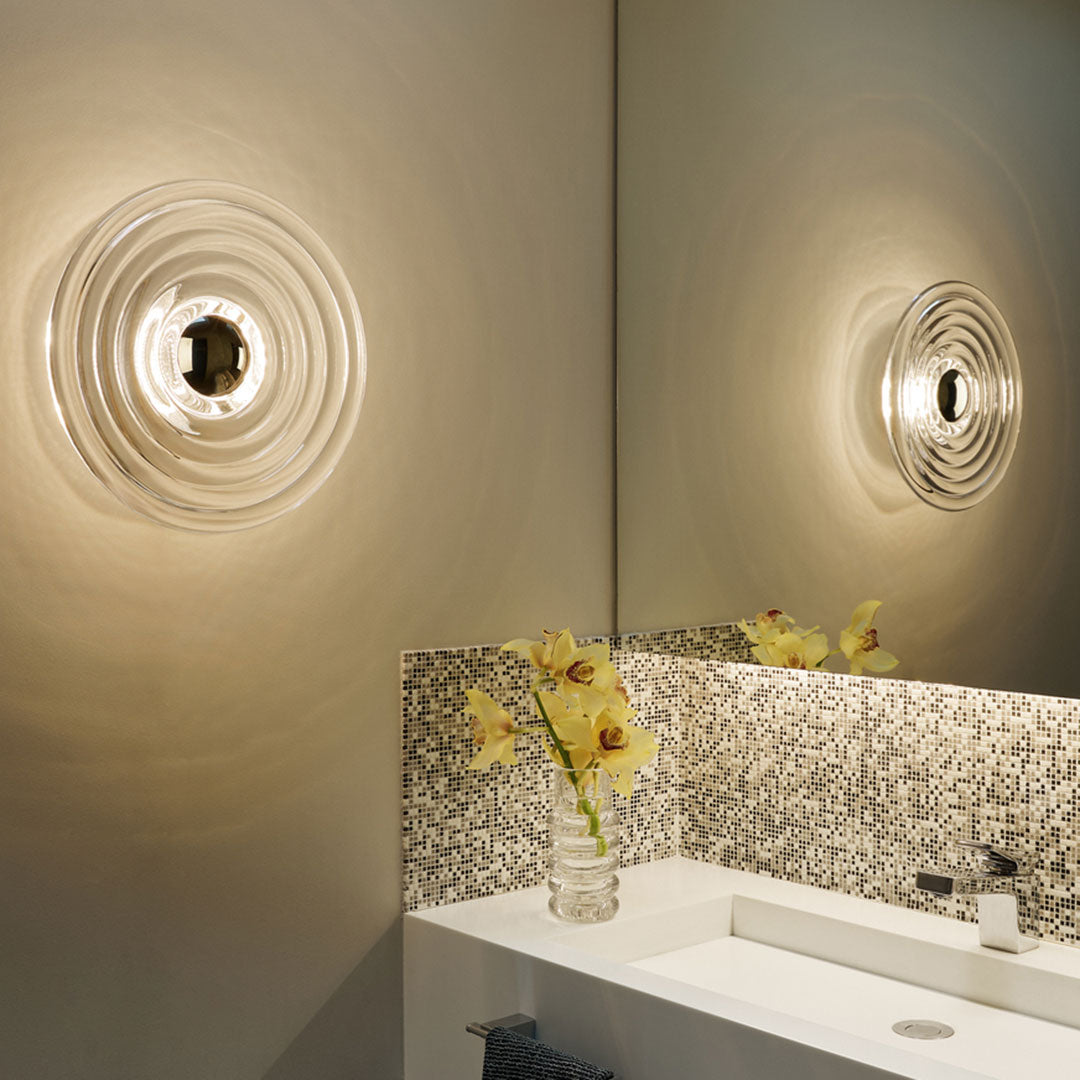 "Transparent round wall light with circular rippled pattern, illuminating a neutral-toned bathroom with speckled mosaic tiles."