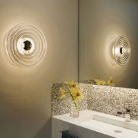 "Transparent round wall light with circular rippled pattern, illuminating a neutral-toned bathroom with speckled mosaic tiles."