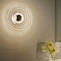 Clear glass wall light with ripple design, complemented by a textured silver pattern and soft yellow floral arrangement.