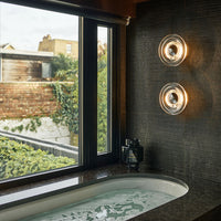 Elegant bathroom scene featuring dark patterned tiles, clear glass light fixtures, and a white soaking tub. Natural greenery outside.