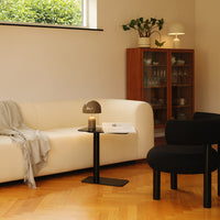 White curved sofa with a gray blanket, black side table, black chair, and warm wooden flooring in a modern setting.
