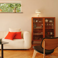 Cozy living room featuring a cream sofa with red pillows, wooden cabinet, glassware, and warm earth-tone accents.