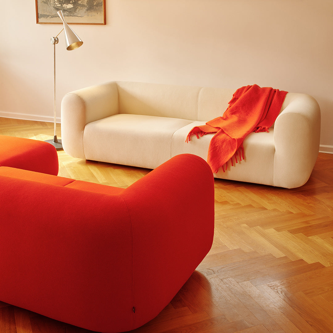 Modern white sofa with a vibrant orange throw, paired with bold red accent chairs, set against warm wooden flooring.