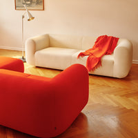 White sofa with soft lines, paired with vibrant orange accent chairs and a cozy red throw blanket. Stylish and contemporary decor.