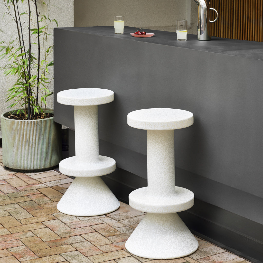 Pair of minimalist white concrete stools with a textured finish, set against a dark gray background and brick flooring.