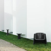 Black textured chair and side tables against a minimalist white wall, set on green grass with a gravel pathway.