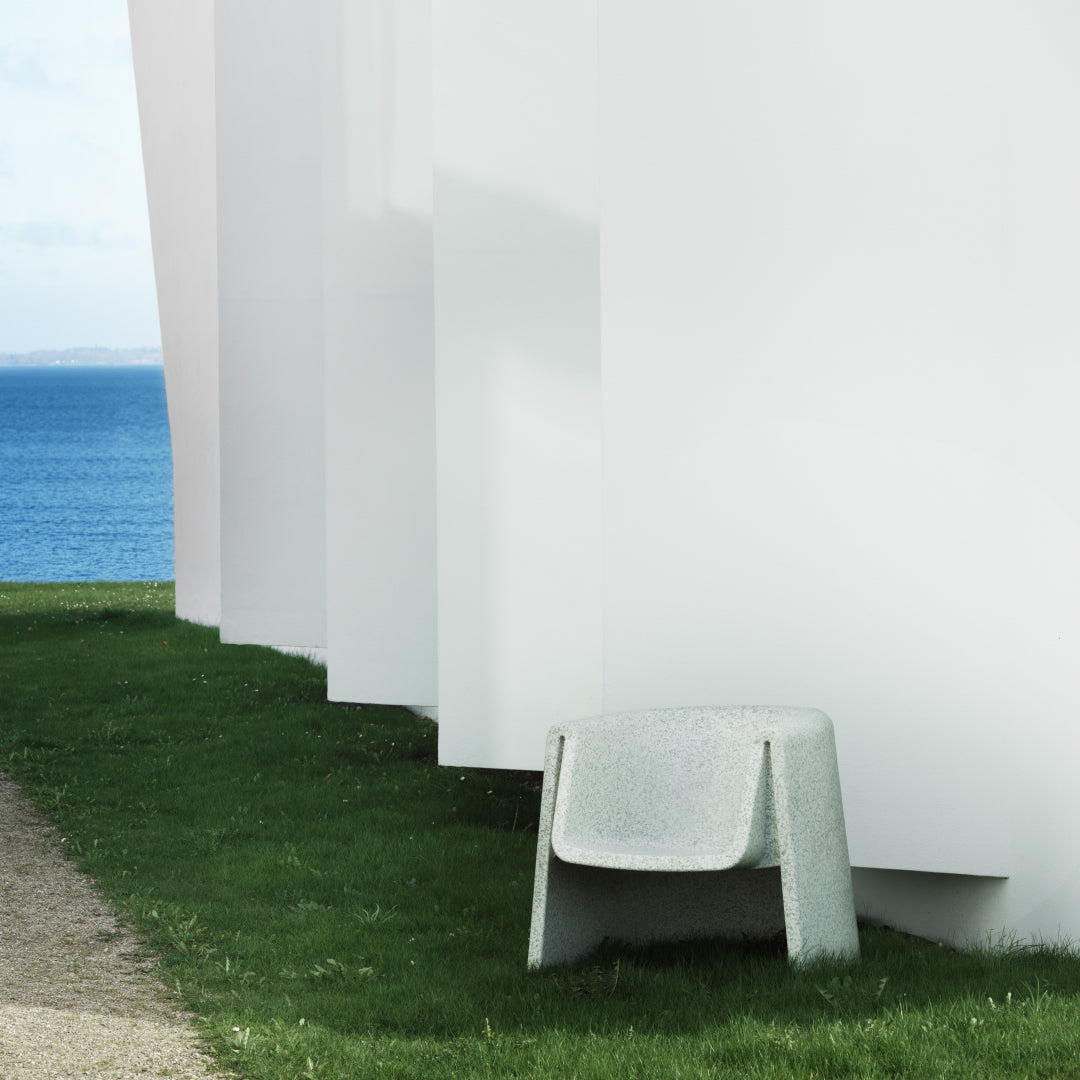Gray chair against white wall, with green grass and blue ocean in the background. Minimalist color scheme.