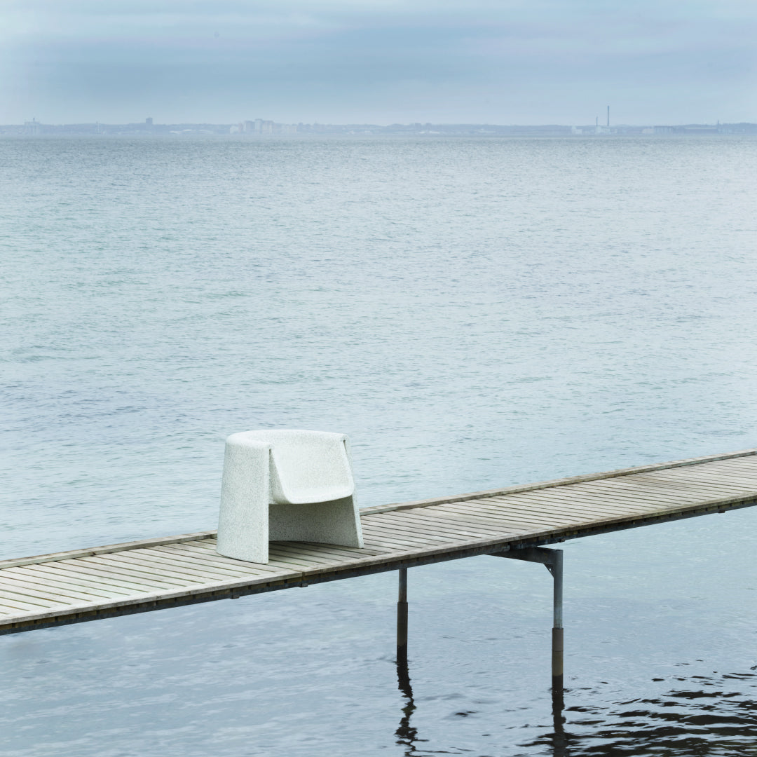 Minimalist white chair on wooden dock, set against a soft blue-gray sea and cloudy sky.