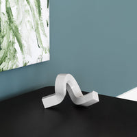 Sleek white decorative sculpture with smooth curves, set against a dark tabletop and backdrop of green abstract art.