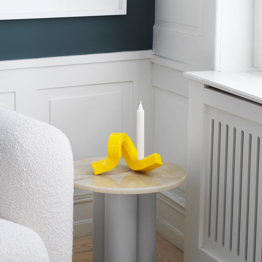 "Yellow curved candle holder on a light wood table, complemented by a white candle and a neutral-colored decor."