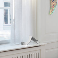 Light, sheer white curtains gently draped near a sleek, silver curtain holder on a light-colored windowsill.