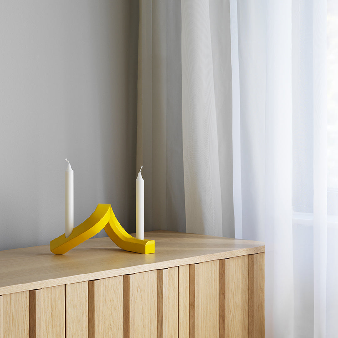 Yellow candle holder with a wavy design, set against a light gray wall and soft white curtains.