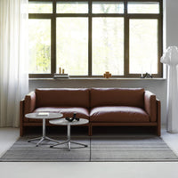 Brown leather sofa with silver accent tables on a gray striped rug, set against a bright, natural light-filled window.