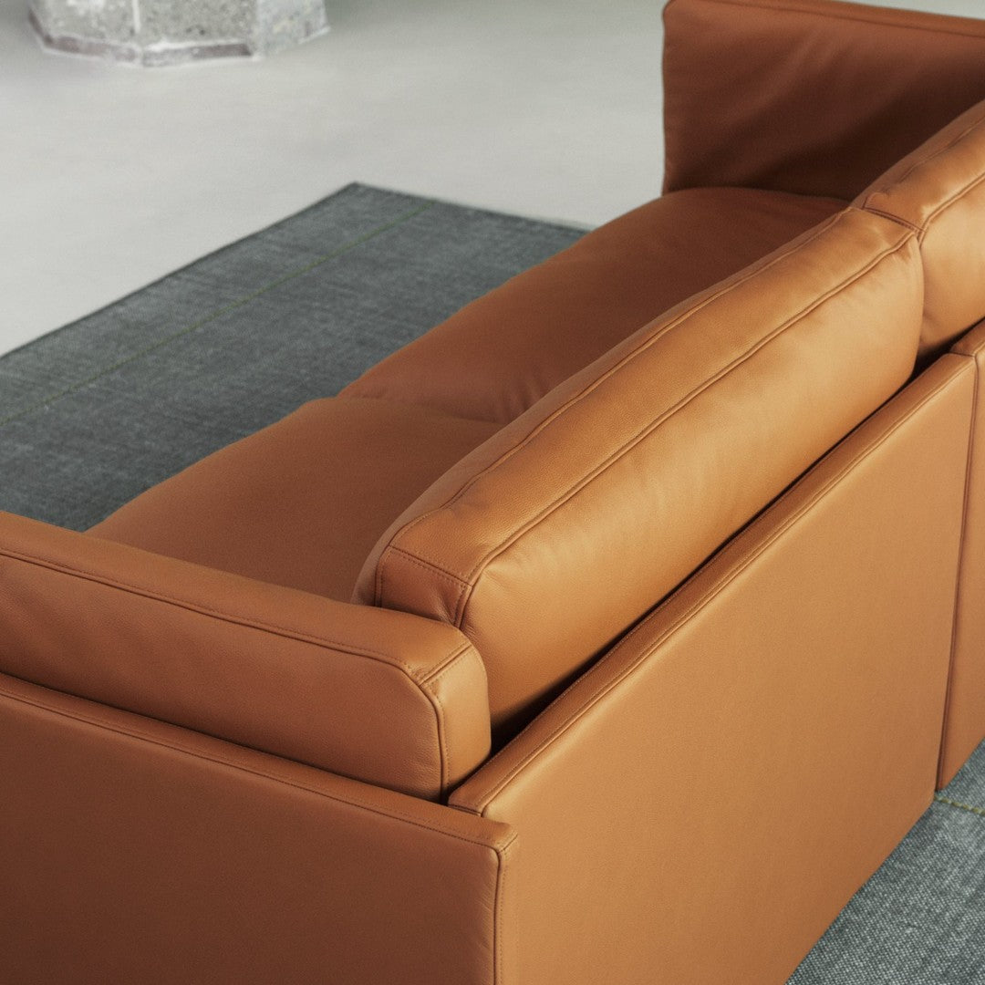 Tan leather sofa with sleek design, featuring smooth surfaces against a gray woven rug with green accents.