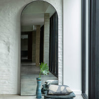 Arched full-length mirror with a sleek black frame, reflecting neutral tones and a textured backdrop.