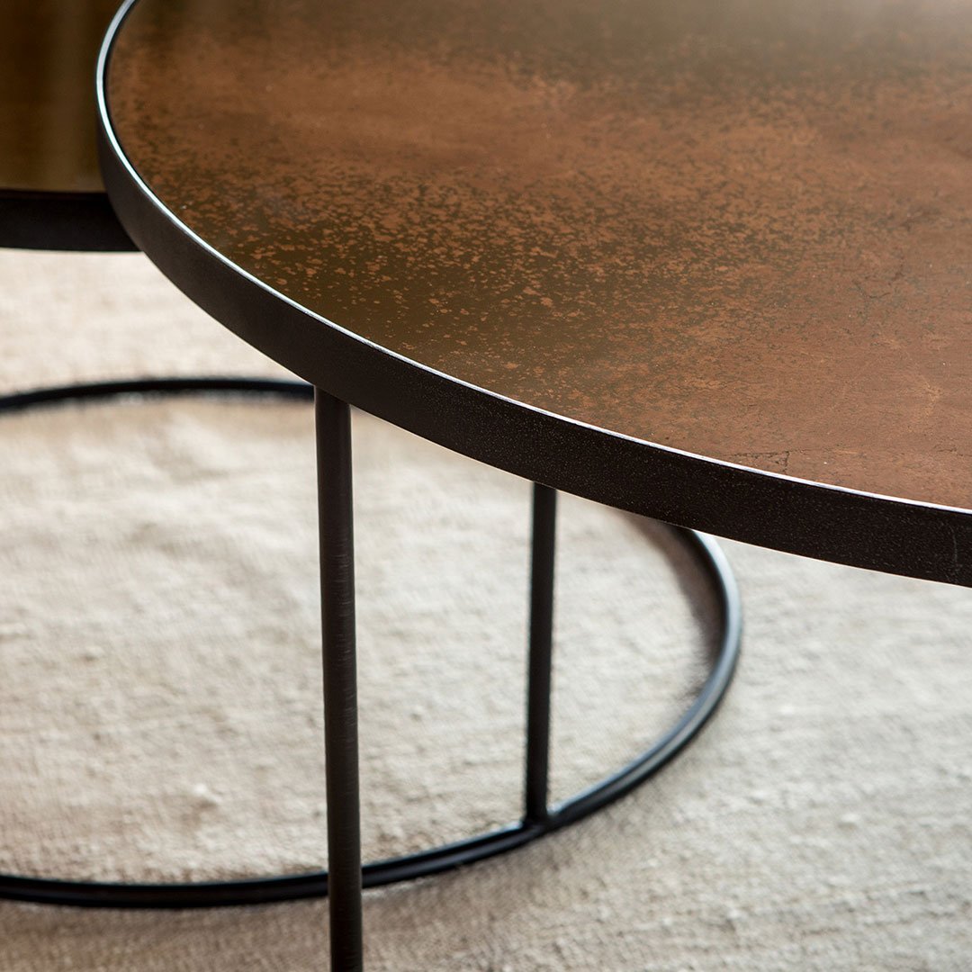 Round coffee table with a textured bronze surface and black metal base, set on a neutral fabric background.