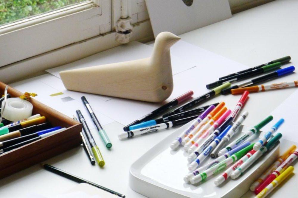 "Colorful assortment of pens and markers on a desk with a minimalist wooden bird figurine."