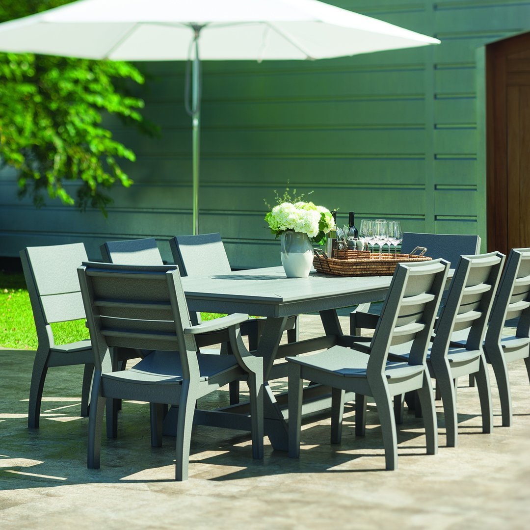 Gray outdoor dining table set with matching chairs, surrounded by greenery and a large white umbrella.