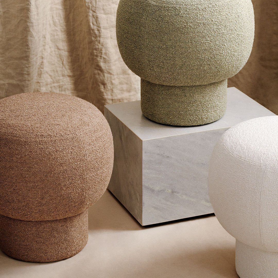 Textured stools in earthy brown, soft green, and crisp white, set on a sleek marble pedestal with a neutral backdrop.
