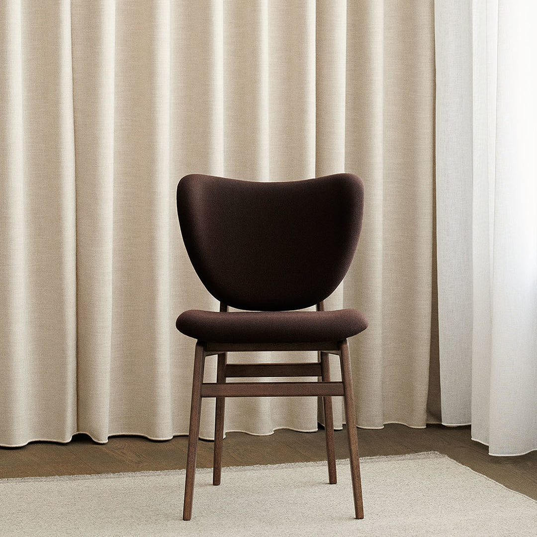 "Elegant brown upholstered chair with curved backrest against soft beige curtains and neutral-toned rug."