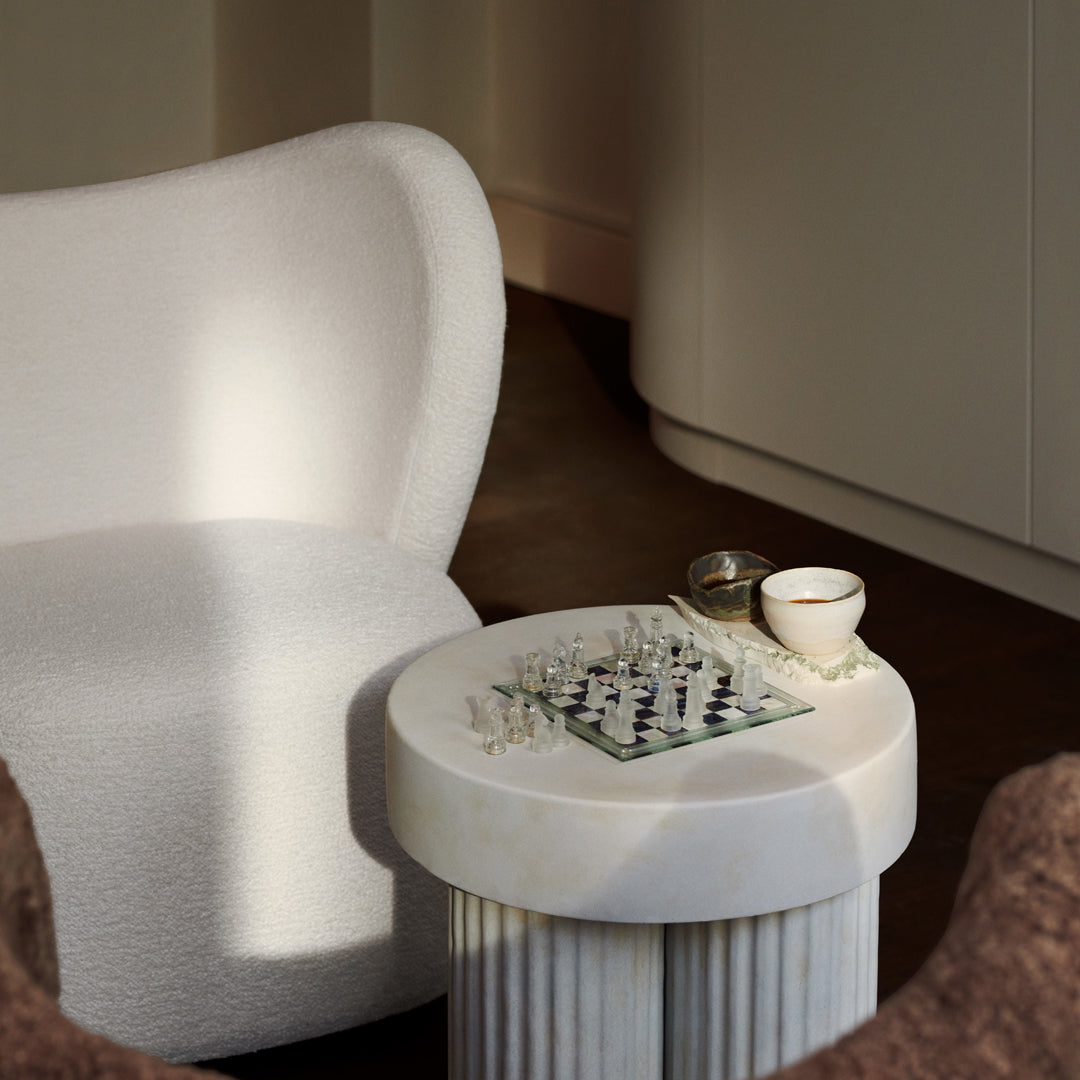 White plush armchair beside a round white column table, featuring a chess set and a beige cup in soft natural lighting.