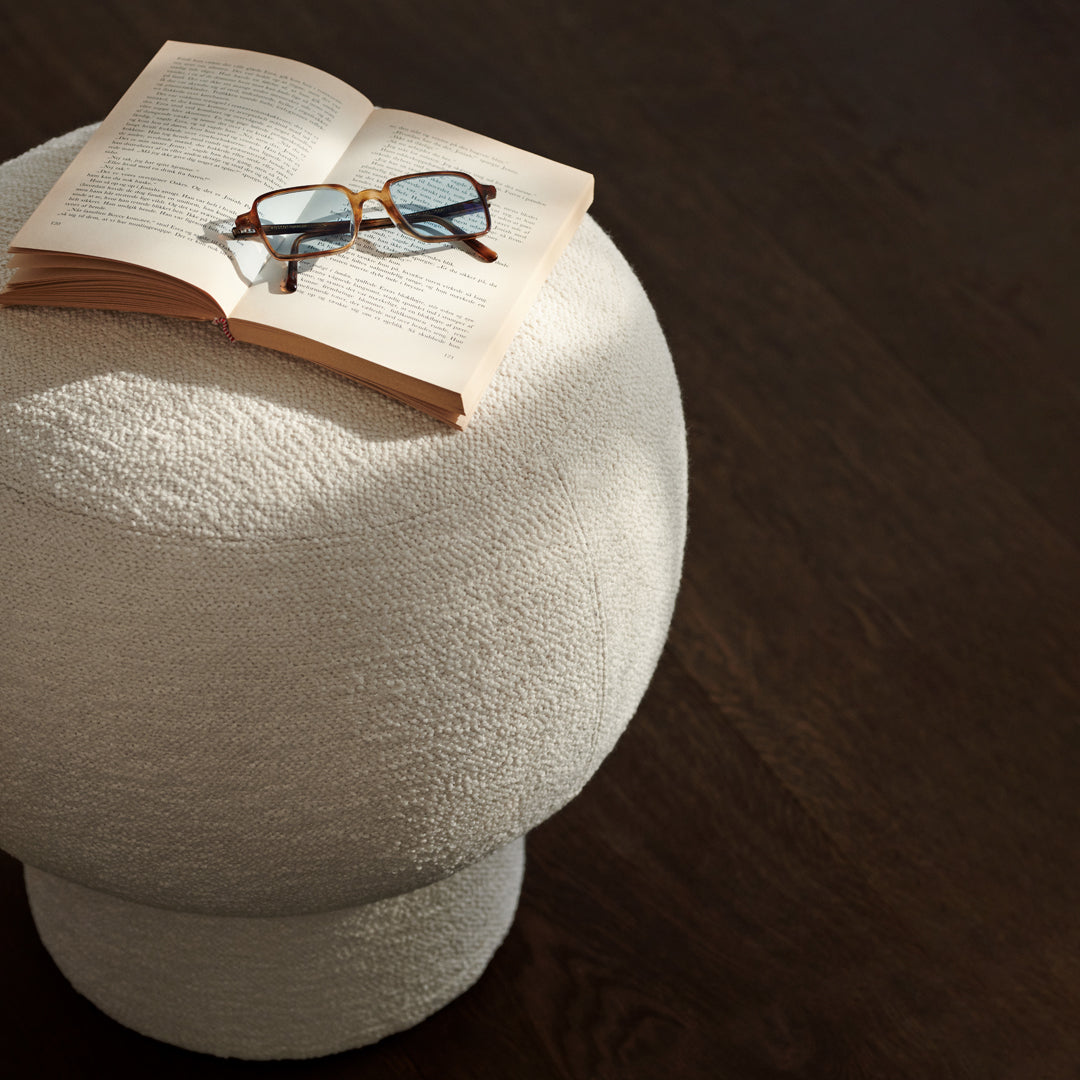 "Round textured white stool on dark wood surface, with an open book and glasses adding a cozy touch."