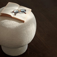 "Round textured white stool on dark wood surface, with an open book and glasses adding a cozy touch."