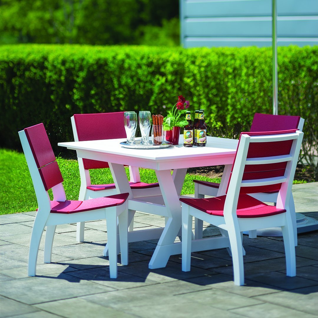 White outdoor table with four pink-accented chairs, set on a stone patio surrounded by lush greenery.