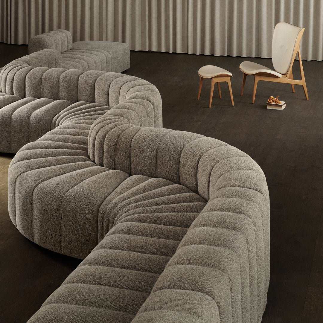 Curved gray sectional sofa with textured, ribbed patterns on a dark wood floor, complemented by light wooden chairs.