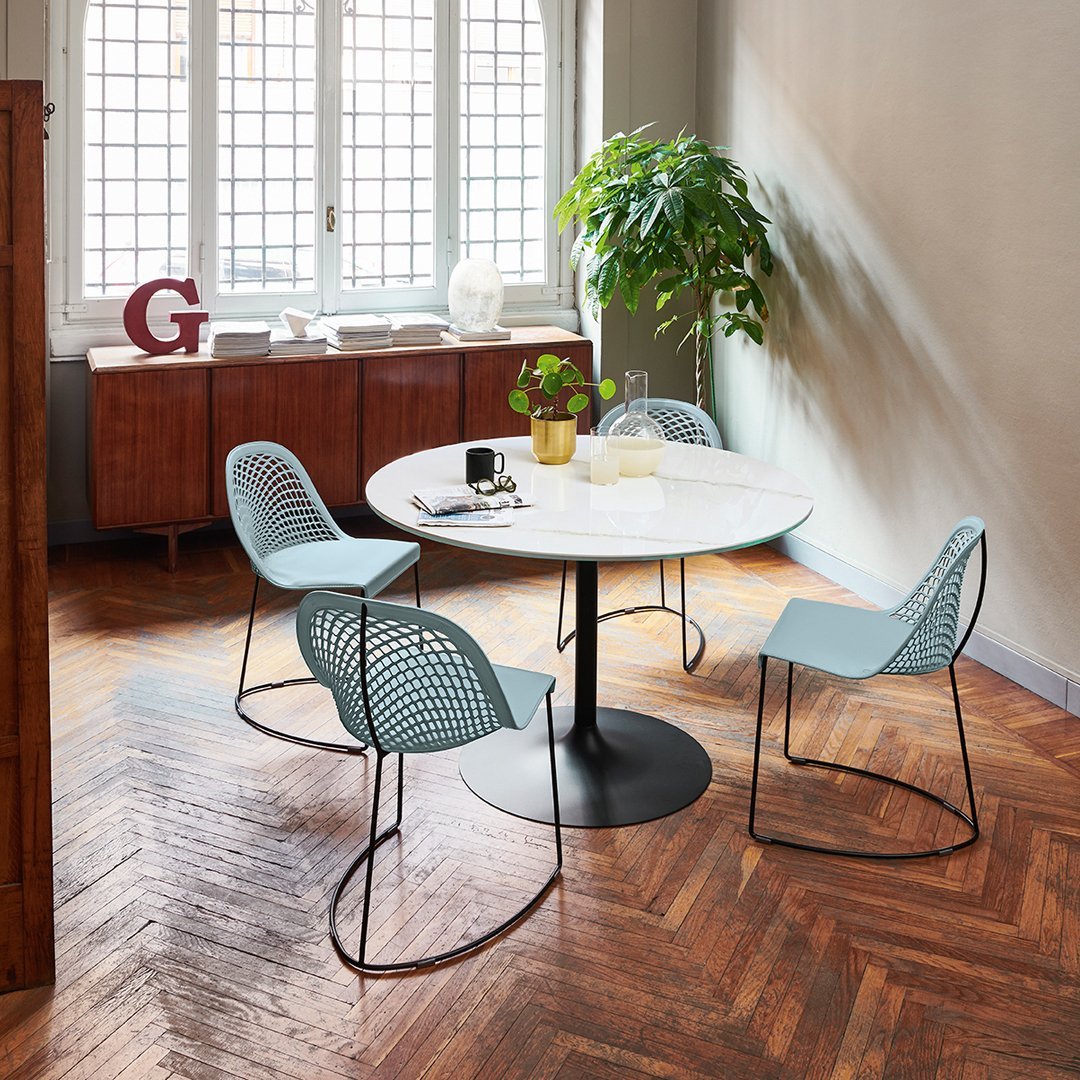 Round white table with light blue woven chairs and warm wooden herringbone flooring, accented by green plant foliage.
