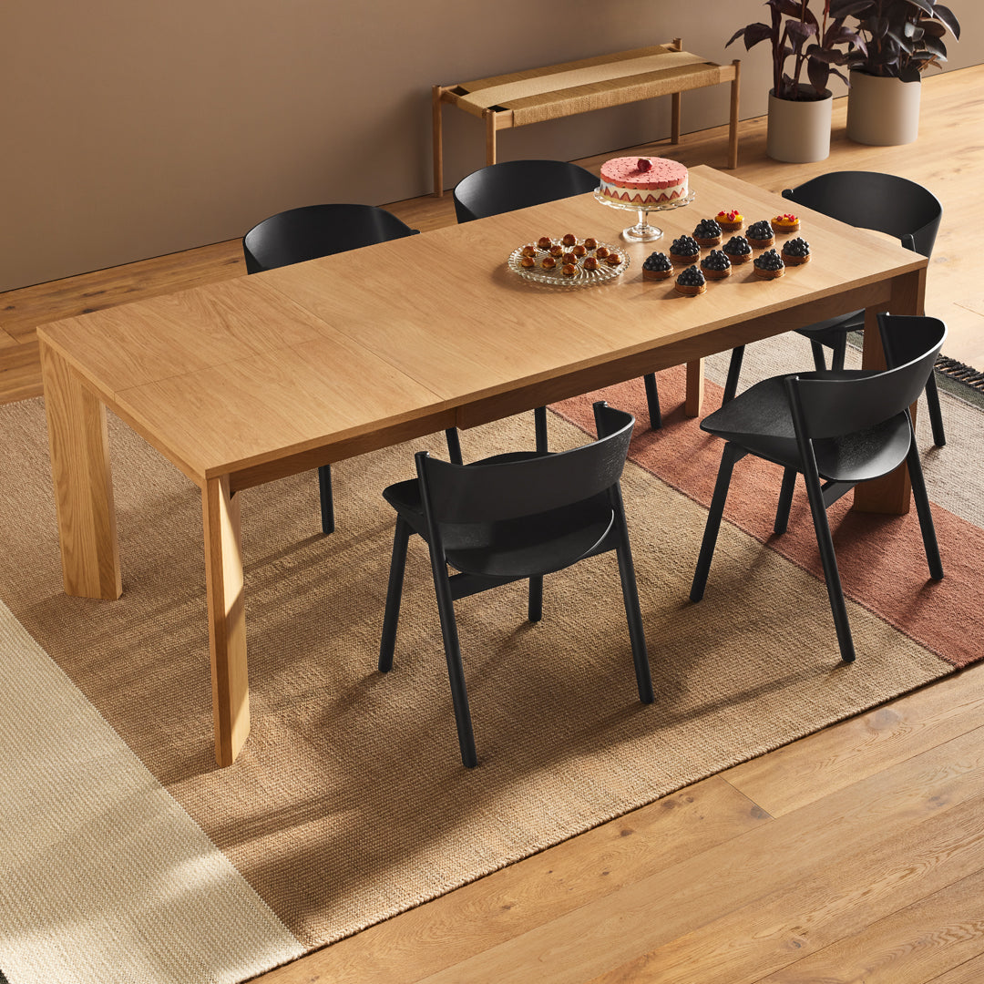 Light wood dining table with black chairs, set on a textured beige rug and surrounded by warm wooden tones.