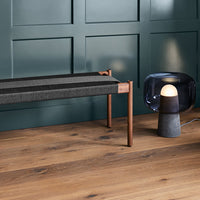 Stylish bench with gray striped upholstery and warm wood legs, set against a deep green wall and rustic wooden floor.