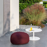 Burgundy round pouf next to a sleek white table, surrounded by lush green plants and neutral stone decor.