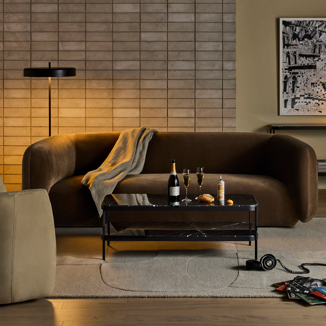 Brown curved couch with a light throw, black coffee table, and neutral-toned rug, set against a textured beige wall.