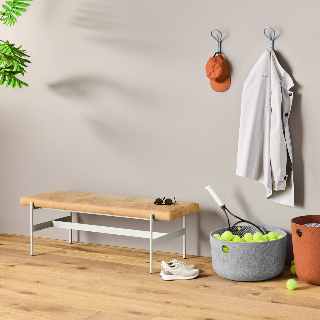 Neutral gray walls with a natural wood bench, white metal frame, orange accents, and a gray fabric basket filled with green tennis balls.