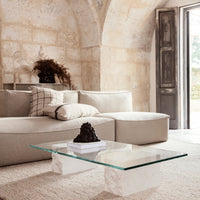 Beige sectional sofa with patterned cushions, minimalist glass coffee table on a textured rug in a light, airy space.