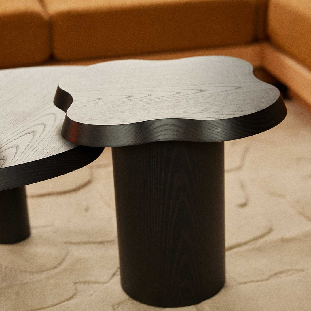 Black wood coffee table with a unique cloud-shaped top and cylindrical base, set against a beige carpet and brown sofa.