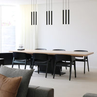 Modern black dining chairs around a light wooden table in a minimalist, neutral-toned room with linear black pendant lights.