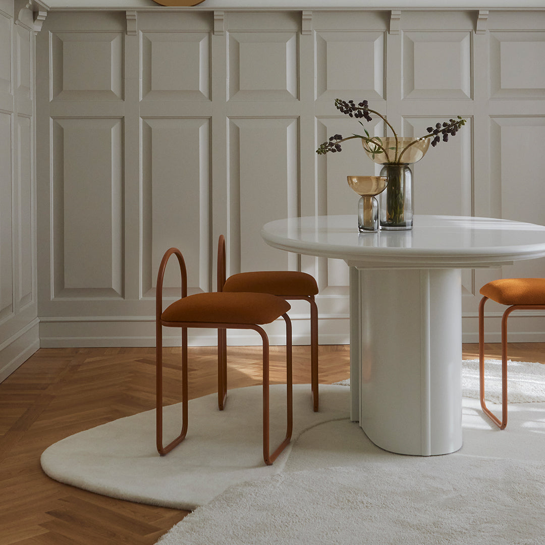Modern dining set featuring orange chairs on a soft white rug, surrounded by neutral beige paneling and wooden flooring.
