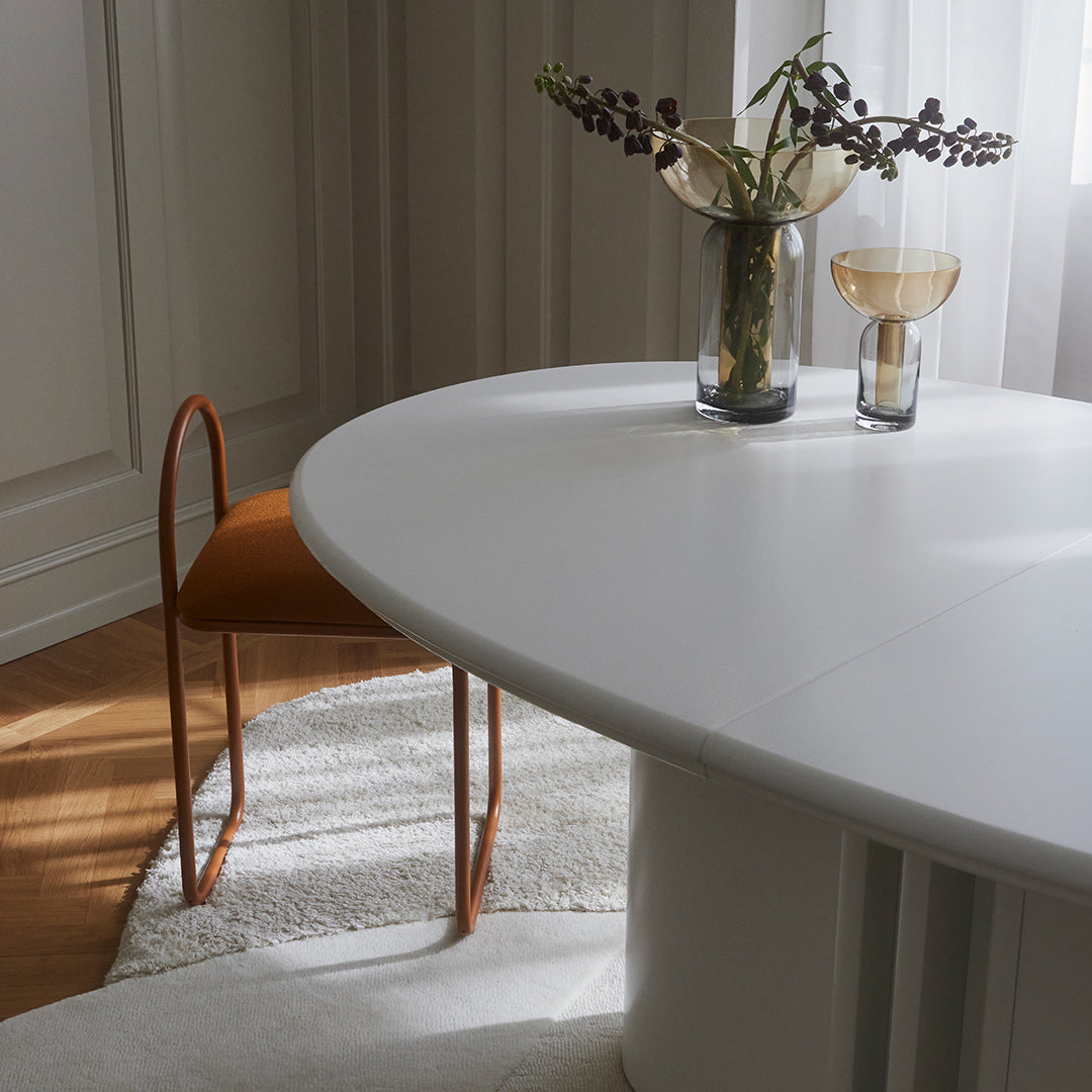 White oval dining table with warm orange chair, neutral textured rug, and elegant glass vases featuring natural decor.