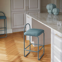 Teal upholstered bar stool with a sleek metal frame, set against light wood flooring and pastel decor accents.