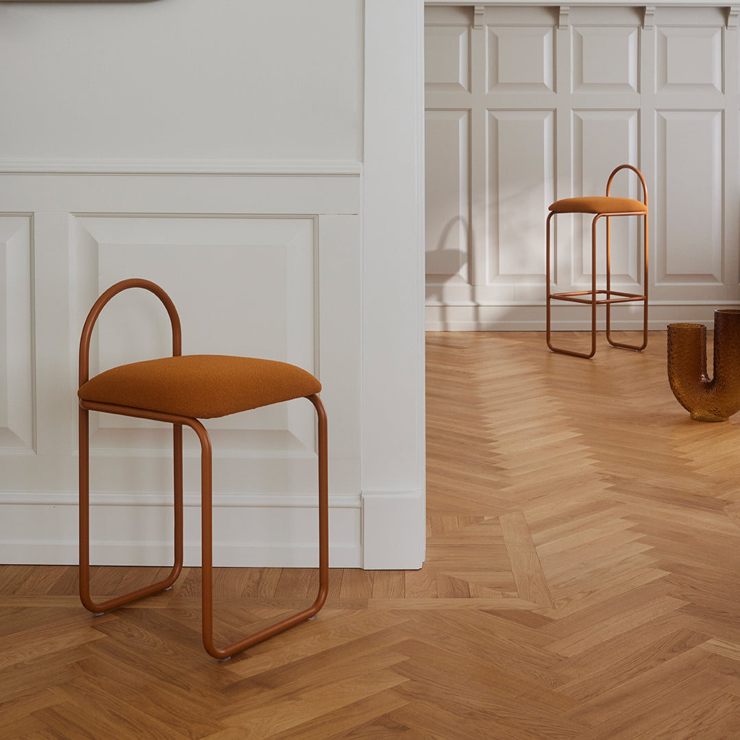 "Two modern stools featuring warm orange upholstery and sleek copper frames on a herringbone wood floor."